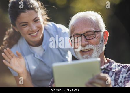 Senior man and young woman caregiver using tablet for video call outdoor in park Stock Photo