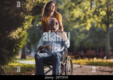 Pretty young woman pushing smiling father in wheelchair in park and having nice time in walk Stock Photo