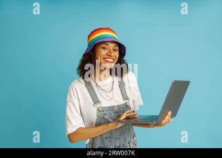 Stylish woman freelancer in rainbow cap working on laptop while standing over blue studio background Stock Photo