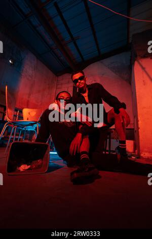 a couple of gay teenagers are sitting together wearing sunglasses and posing naughty in a warehouse with a red light at night Stock Photo
