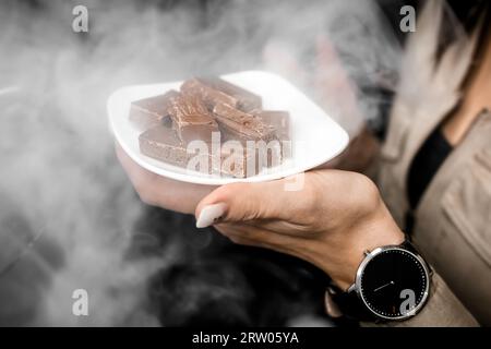 The girl attracts attention by holding in her hand a white saucer with pieces of chocolate in the smoke from the hookah, nightlife concept. Stock Photo