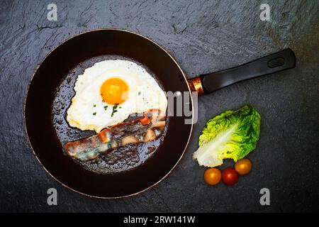Fried egg with a strip of bacon in a pan. On the right are 3 tomatoes and a leaf of green lettuce.Photographed from a bird's eye view in front of Stock Photo