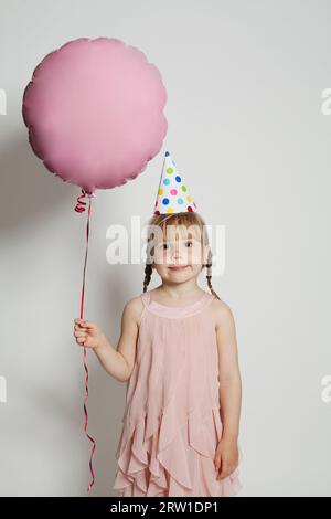 Positive small kid dressed in pink festive clothes, carries air balloons, celebrates holiday. Childhood, birthday party and celebration concept Stock Photo