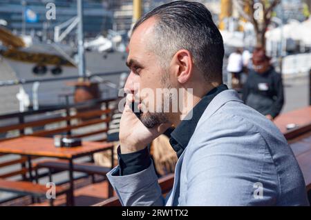 Profile view of elegant business caucasian man with beard in gray jacket sitting outside the restaurant on call talking on phone Stock Photo