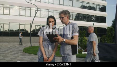 Two doctors stands outdoor and discuss patient treatment. Health care specialists look at client tests results using tablet. Woman helps colleague with making diagnosis. Medical staff work outside. Stock Photo