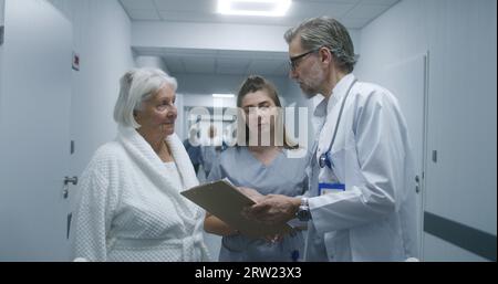 Mature doctor walks the clinic corridor, comes to colleague and elderly patient. Physician shows test results on papers to nurse and woman. Medical staff and patients in hospital hallway. Stock Photo