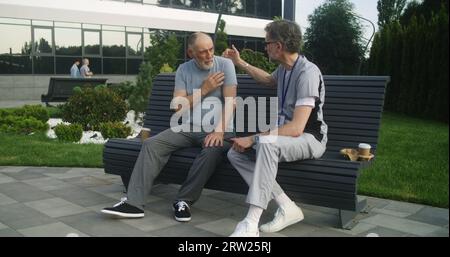 Health care specialist sits with aged patient on bench and discusses his treatment. Elderly man complains of shoulders pain to mature doctor. Professional medic in uniform consults client outdoor. Stock Photo