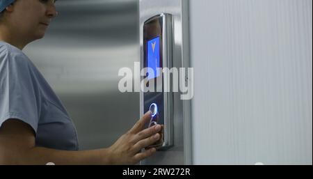 Close up shot of nurse in uniform coming to lift door, pressing call button. Healthcare specialist at work in modern clinic. Medical worker and elderly patient wait elevator in hallway. Stock Photo