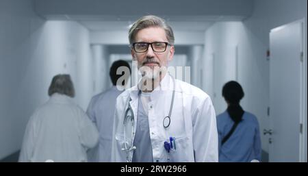 Mature doctor stands in modern clinic corridor. Professional medic puts on glasses, smiles, looks at camera. Medical staff and patients in background in hospital or medical center hallway. Portrait. Stock Photo