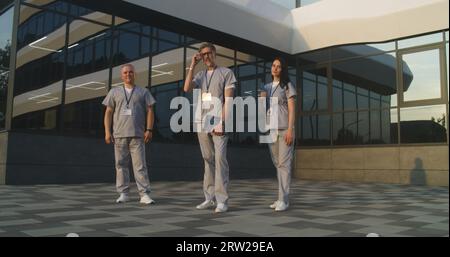 Diverse doctors in uniform stand outdoors near the hospital building. Professional medics and nurse look at camera. Medical staff of modern clinic or medical center. Concept of healthcare system. Stock Photo