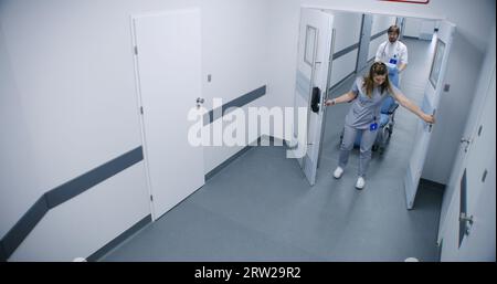 Clinic corridor: Doctors, professional medics and patients walking. Male nurse pushes wheelchair. Elderly man goes to his hospital room. Medical staff work in modern medical facility. High angle. Stock Photo