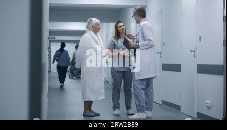 Nurse and elderly woman walk the clinic corridor after procedures. Male doctor comes to colleague and patient, shows tests results using digital tablet. Medical staff and patients in hospital hallway. Stock Photo