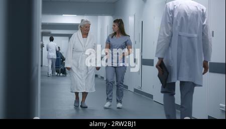 Nurse and elderly woman walk the clinic corridor after procedures. Male doctor comes to colleague and patient, shows tests results using digital tablet. Medical staff and patients in hospital hallway. Stock Photo