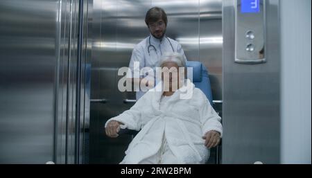 Doctor moves elderly woman in transport wheelchair from elevator. Medic ...