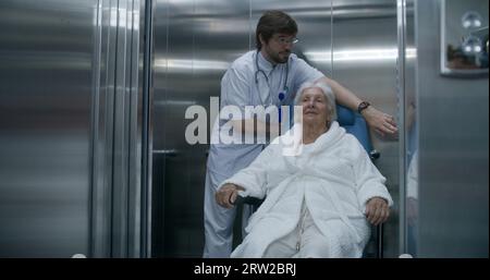 Doctor moves elderly woman in transport wheelchair from elevator. Medic ...