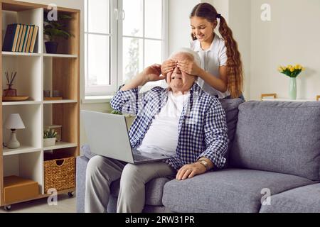 Cute smiling girl covering eyes with hands of senior grandfather while playing together at home. Stock Photo
