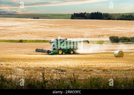 Rocky View County Alberta Canada, September 11 2023: A John Deere Combine harvesting a crop with a full hopper of grain passing a round hay bale on th Stock Photo