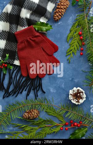 Fringed black and white scarf and red gloves on blue background with winter greenery, berries and cones in flat lay composition. Winter concept. Stock Photo