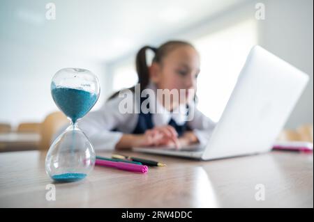 Caucasian girl sits at a desk at school and studies at a laptop. Hourglass on the table. Stock Photo