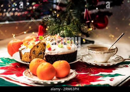 Christmas stollen with glaze, candied fruits and nuts on a saucer with a cup of tea, tangerines near a small Christmas tree. A festively set table for Stock Photo