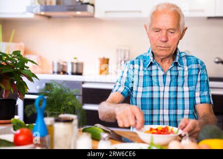 Elderly man cuts vegetables for salad at the table in the kitchen Stock Photo