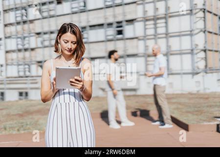 Female construction engineer reviewing blueprint on tablet. Her male partners discussing in the back. Copy space. Stock Photo