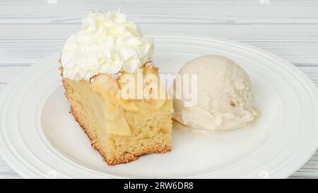 Slice of fresh baked apple cake with biscuit base and sliced apples and whipped cream topping served with vanilla ice cream close up on a plate on whi Stock Photo