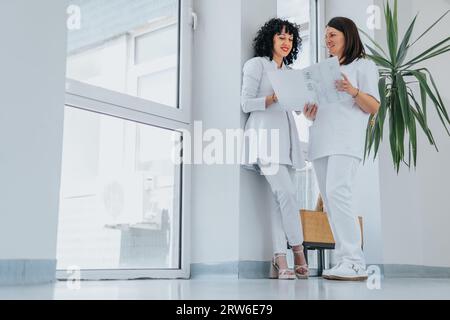 Female doctors standing in a hall at the hospital, discussing positive patients results and smiling. Stock Photo