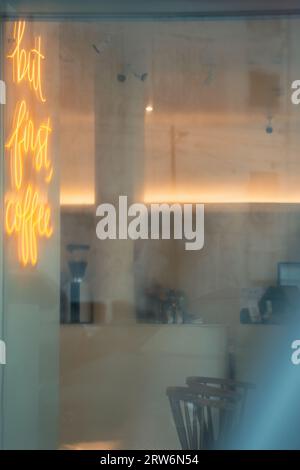 An orange neon script sign glows inside a cozy, blurred coffee shop interior viewed through a window with reflections. Stock Photo