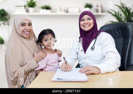 Portrait of smiling young muslim doctor wearing hijab checking girl child at modern clinic. Friendly female pediatrician examine young kid patient wit Stock Photo