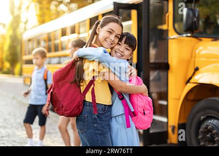 Portrait Of Two Happy Girls Embracing While Standing Outdoors Near School Bus Stock Photo