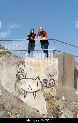Two people standing behind railings above graffitied concrete wall with 'Eat The Rich' message Stock Photo