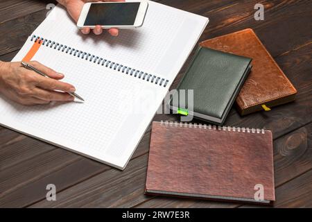 Hands hold pen and smartphone, opened notebook. On table  calculator and notebooks. Top view. Dark wood background Stock Photo