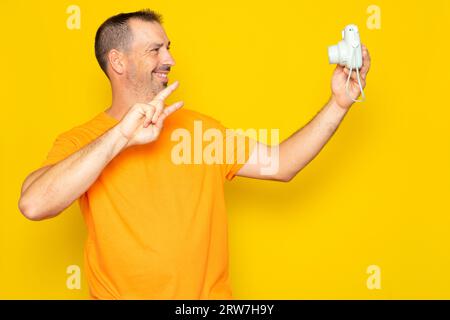 Bearded Hispanic man in his 40s taking a selfie with an instant camera, isolated on yellow studio background. Stock Photo