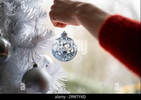 Closeup view of female hand in red sweater hanging shiny blue holiday bauble on a white christmas tree. Stock Photo