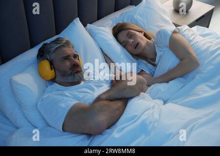 Irritated man with headphones lying near his snoring wife in bed at home Stock Photo