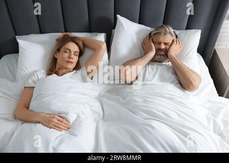 Irritated man with headphones lying near his snoring wife in bed at home Stock Photo