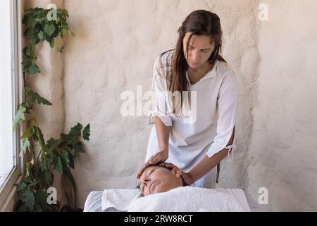 Beautiful young therapist performing massage in spa salon. Front view portrait of beautician doing facial massage to relaxed female client. Facial bea Stock Photo