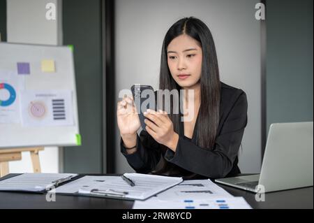 A focused and thoughtful Asian businesswoman is reading messages on her smartphone with a serious face while sitting at her desk in the office. Stock Photo