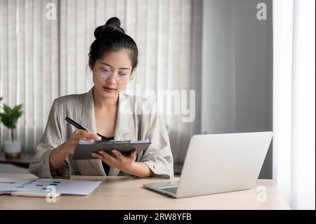 A focused and professional millennial Asian businesswoman or female manager in a casual suit examines business documents on a clipboard at her desk. Stock Photo