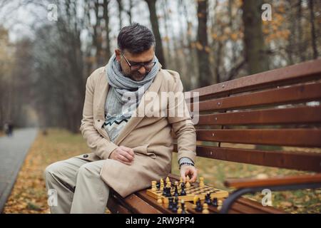 An elderly man plays chess on a bench in an autumn park. Stock Photo