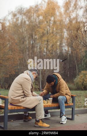 A white senior and a Hindu young man are playing a game of chess on a bench in an autumn park. Generational friendship concept Stock Photo