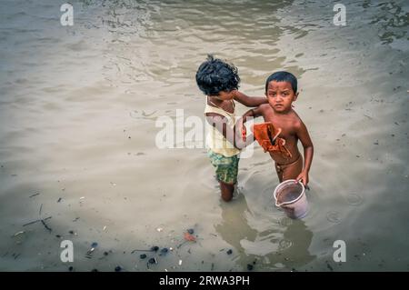 Sunderban, Bangladesh, circa July 2012: Small native children wash in river and boy holds bucket and frowns to photocamera in Sunderban, Bangladesh. Stock Photo