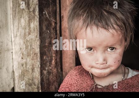 Dolpo, Nepal, circa May 2012: Small girl with brown glittering eyes dressed in red sweater looks curiously up to photocamera in Dolpo, Nepal. Stock Photo