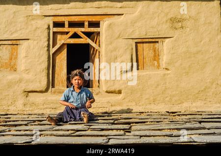 Beni, Nepal, circa May 2012: Small girl in blue shirt and two ponytails sits on ground in front of their house in Beni, Nepal. Documentary editorial Stock Photo