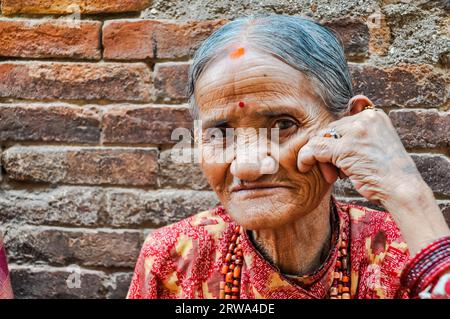 Bhaktapur, Nepal, circa June 2012: Old grey-haired woman with wrinkled face with necklace made of beads around her neck holds her hand on cheek in Stock Photo