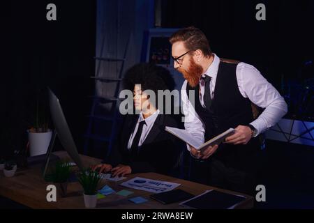 african american woman typing on computer near bearded businessman with notebook in office at night Stock Photo