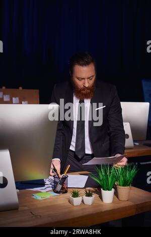 bearded and concentrated businessman working with documents near computer at night in office Stock Photo