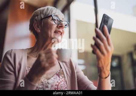 Mature woman is sitting in cafe and relaxing. She is using smartphone. Stock Photo