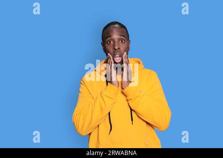 African american man in yellow hoodie, hands on cheeks. Reaction to something amazing. Isolated on blue background. OMG concept. Stock Photo